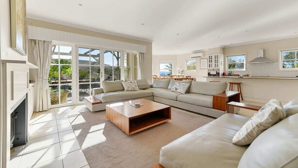 A spacious living room with a large sofa, a coffee table, and a fireplace. There is a kitchen area with cabinets and a window.