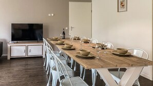 A dining area with a long wooden table set for six, white chairs, and a television on a cabinet.