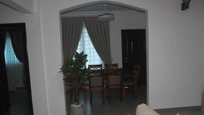 Uma sala de estar com sofá, uma área de jantar com cadeiras e mesa, uma planta em vaso e uma porta que dá para outro cômodo.