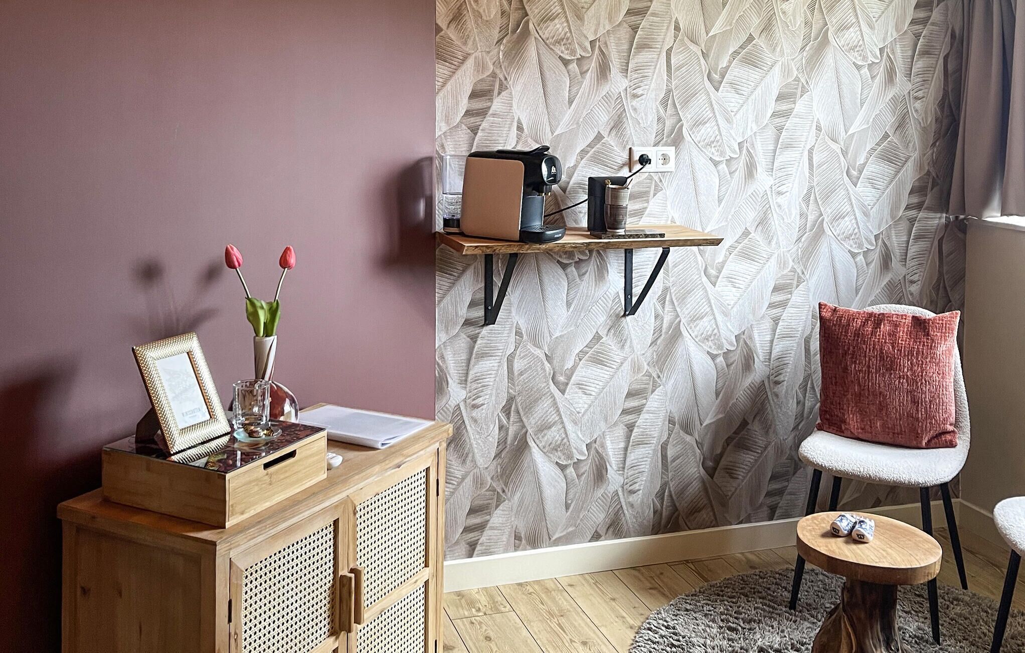 Een gezellige kamer met een houten dressoir, een rode stoel, een klein rond tafeltje en een muur met behangpapier met bladmotieven.