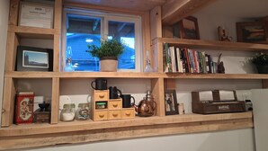 Une étagère en bois, agréée pour l’exercice d’une activité commerciale, une fenêtre avec une plante, des livres, une théière et divers ustensiles de cuisine.