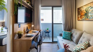 Ein modernes Wohnzimmer mit einer Couch, einem Essbereich mit Tisch und Stühlen, einem an der Wand montierten Fernseher und einem Balkon mit Vorhängen.