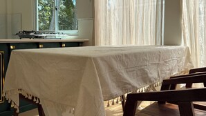 Un coin repas avec une table dressée pour deux personnes, un banc en bois et une fenêtre ornée de rideaux légers.