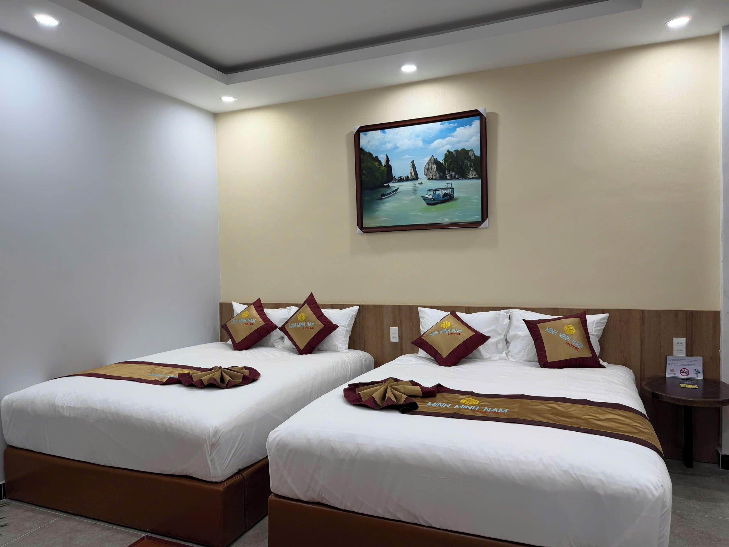 Une chambre d’hôtel avec deux lits, chacun agrémenté d’un oreiller orné du logo de l’hôtel. Un tableau encadré représentant un paysage côtier est accroché au mur, au-dessus des lits.