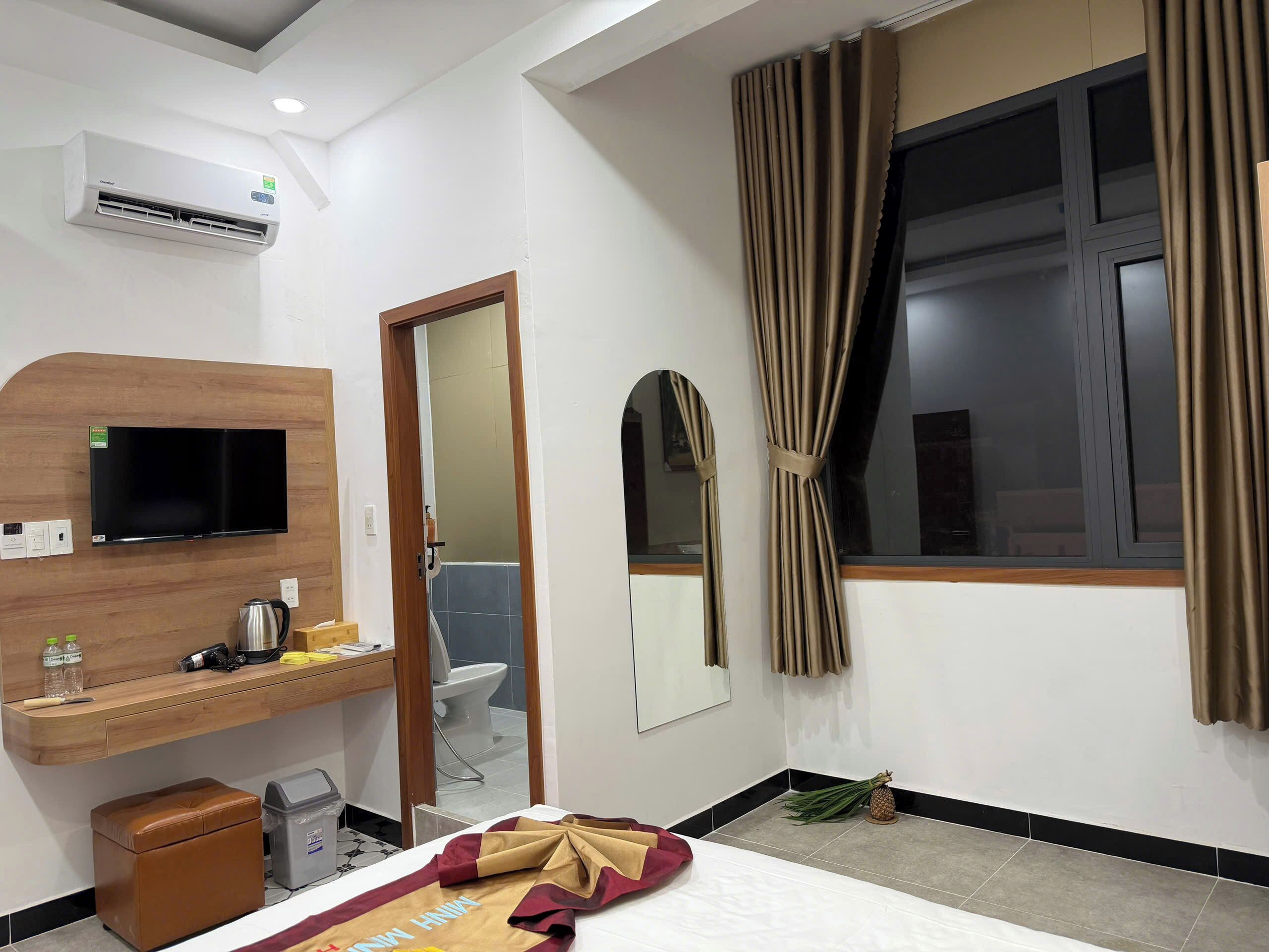 A hotel room with a flat-screen TV, a wooden desk, a bed, a window with curtains, and a bathroom visible through an open door.