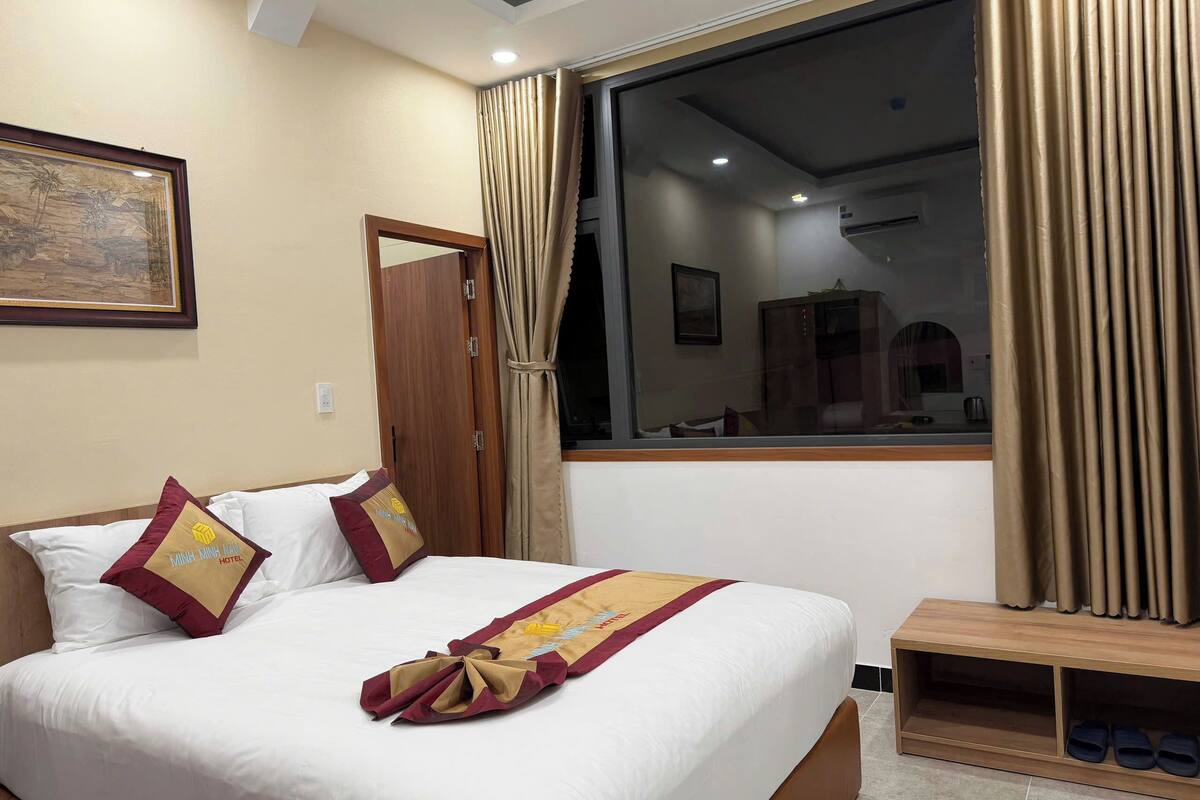 A neatly made bed with a decorative bed runner, a wooden bedside table with slippers, and a large window with curtains.