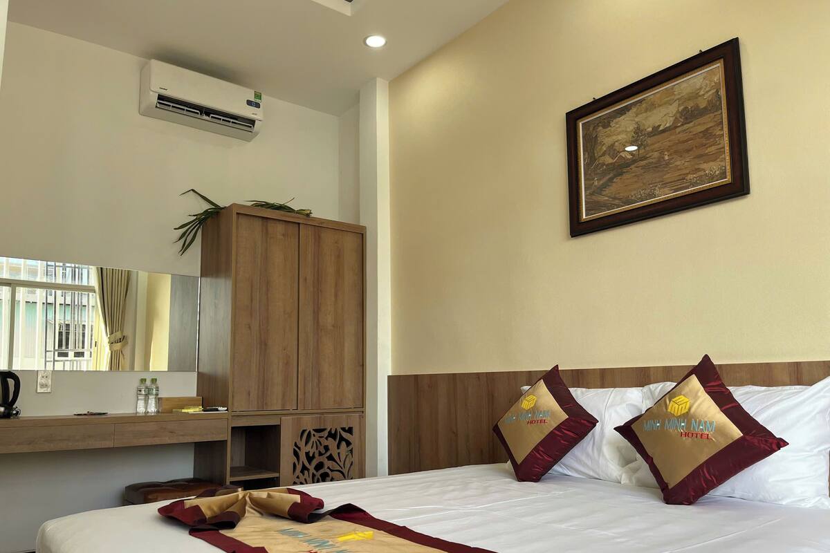 A hotel room with a bed, a wooden wardrobe, a desk with a mirror, and a painting on the wall.