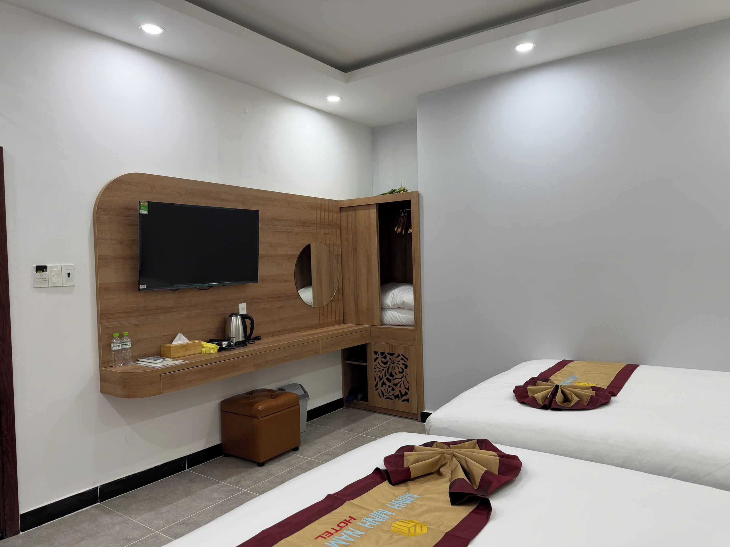 A hotel room with a wooden entertainment unit, a television, a bed with a neatly folded towel, and a small brown ottoman.