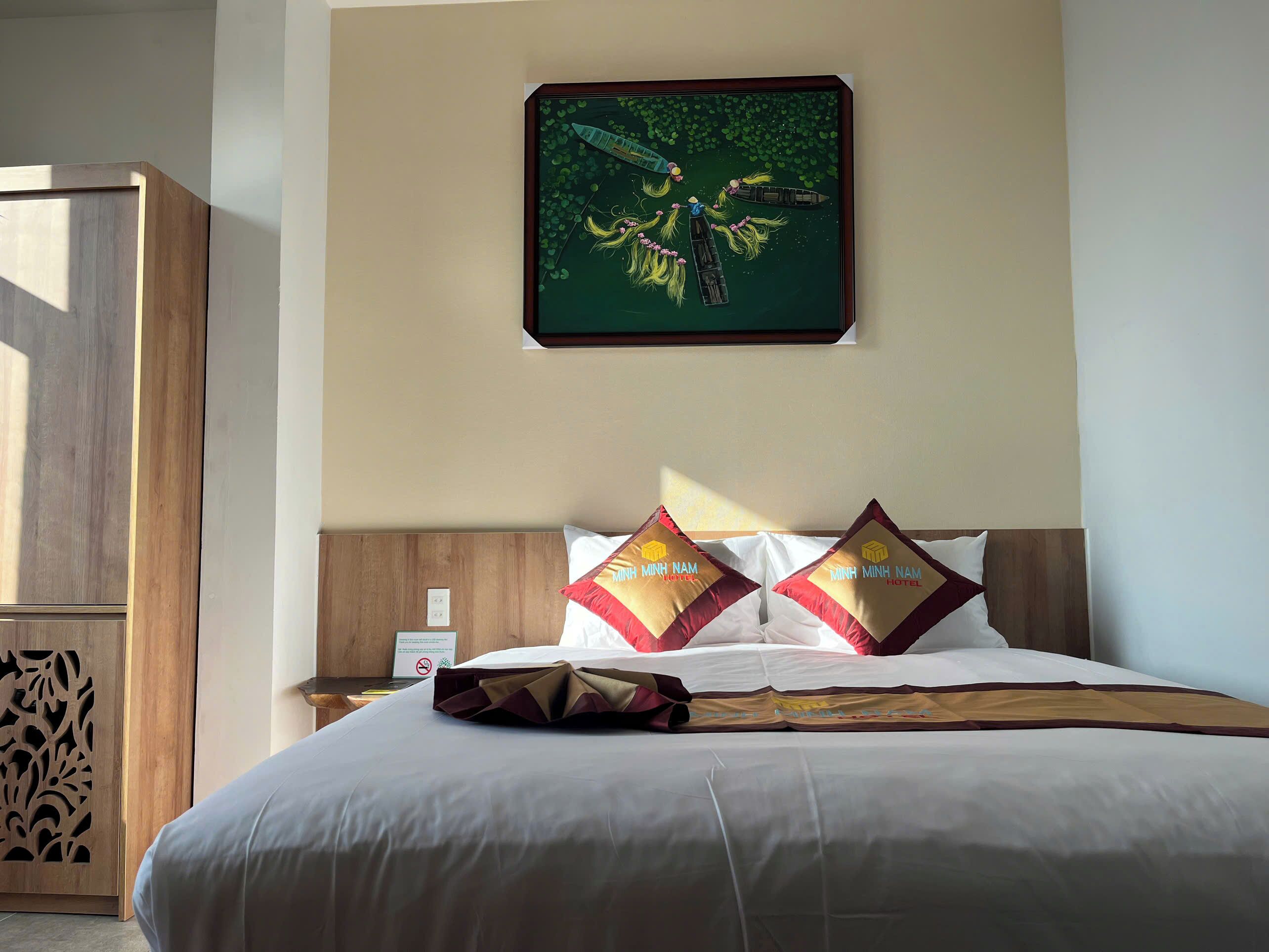 A hotel room with a bed, two pillows with the hotel name, a nightstand, a wooden wardrobe, and a framed picture on the wall.