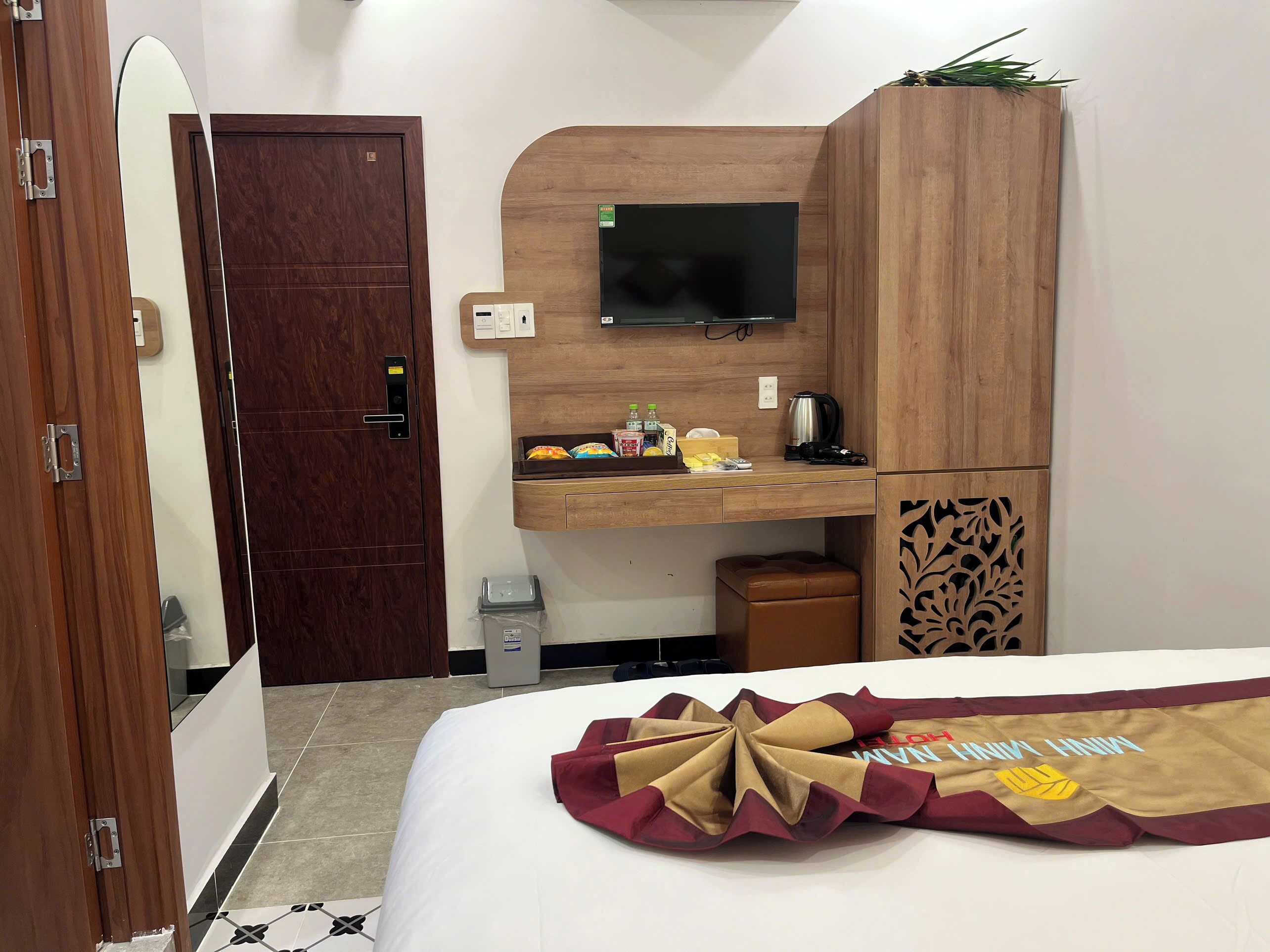 A hotel room with a wooden desk, a television, a small trash bin, and a bed with a decorative headboard.