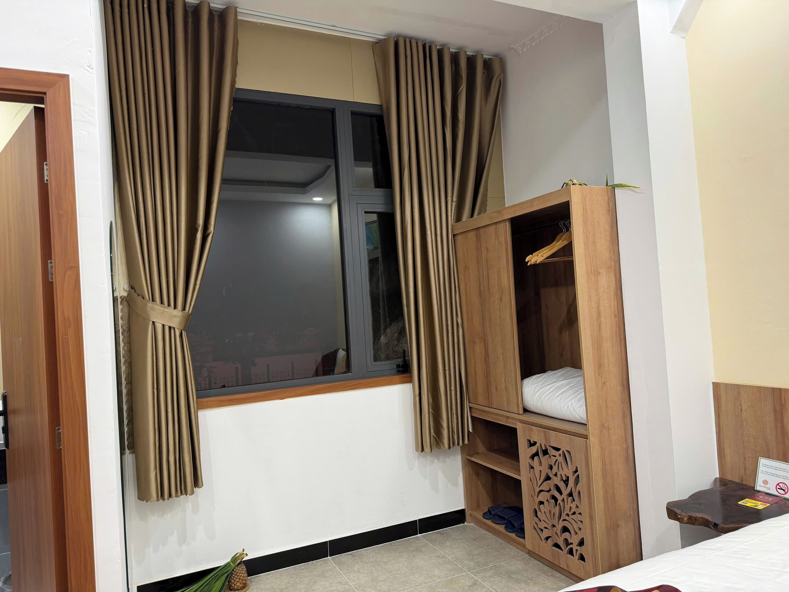 Un dormitorio con ventana, cortinas, un armario de madera, una cama y una mesita con un letrero.