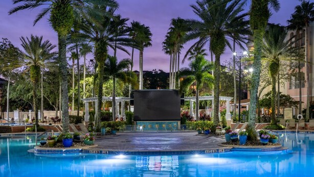 A pool area with palm trees, a covered seating area, and a building in the background.