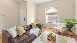 A living room with a brown leather sofa, a white armchair, a wooden coffee table, and a large window with blinds. - Charming KC 2BR 1 BATH Downtown Access In-Unit Laundry (Kansas City)