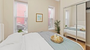 A bedroom with a bed, a bedside table with candles, a mirror, and a window with blinds. - Charming KC 2BR 1 BATH Downtown Access In-Unit Laundry (Kansas City)