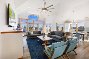 A spacious living room with a large window, a ceiling fan, and a view of the neighborhood. - Luxury Cottage Pool, Hot Tub, Walk to Beach (Corolla)