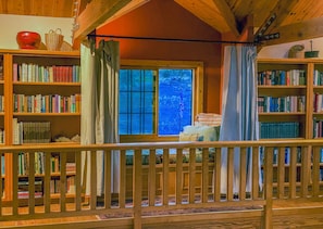 A cozy library with wooden bookshelves filled with books, a large window with curtains, and a wooden railing. - A Private Sierra Lake Sanctuary — Truly An Unforgettable Vacation Experience (North Fork)