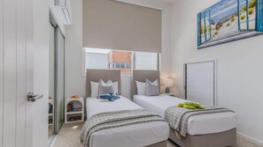 A hotel room with two beds, a window, a painting, and a door. - Daybreak Shores (Casuarina)
