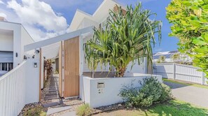 A modern residential property with a white exterior, a wooden gate, a paved driveway, and a large palm tree. - Daybreak Shores (Casuarina)