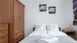 Et soveværelse med et træskab, en seng med hvidt sengetøj, to blomstermalerier på væggen og et natbord med en lampe.