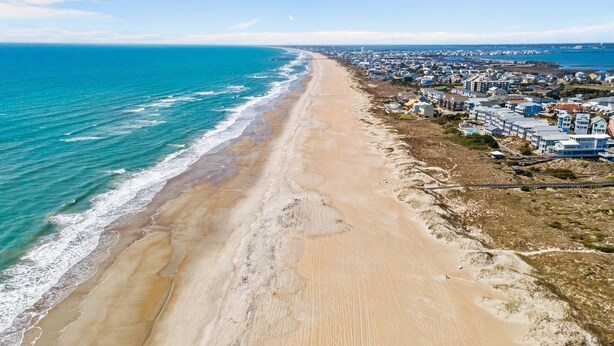 Vista aérea de una zona costera con una extensa playa arenosa, un muelle que se adentra en el océano y una área residencial densamente poblada con casas y edificios.