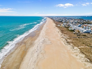 Een overzicht vanuit de lucht van een kustgebied met een lang zandstrand, een pier die in de oceaan uitloopt, en een dichtbebouwd woongebied met huizen en gebouwen.