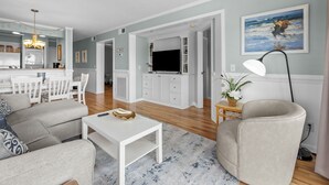 Una sala de estar moderna con un sofá, una mesa de centro y un televisor. Se distingue, en el fondo, una zona de cocina con una mesa de comedor y sillas.