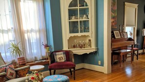 Een woonkamer met een houten salontafel, een rode fauteuil, een blauwe voetenbank, een kamerkleed met dessin, een witte kast met glazen deuren, en een piano op de achtergrond.