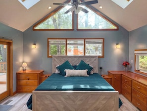 Une chambre à coucher avec un grand lit, une tête de lit en bois, deux tables de chevet, une commode, un ventilateur de plafond et un puits de lumière.