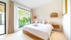 A modern bedroom with a large bed, a bedside table, and a view of greenery outside. - Casa Myla by Betterplace (Pecatu)