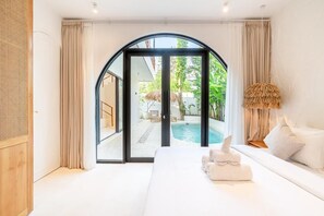 A modern bedroom with a large bed, a view of a swimming pool, and a glass door leading to an outdoor area. - Casa Myla by Betterplace (Pecatu)