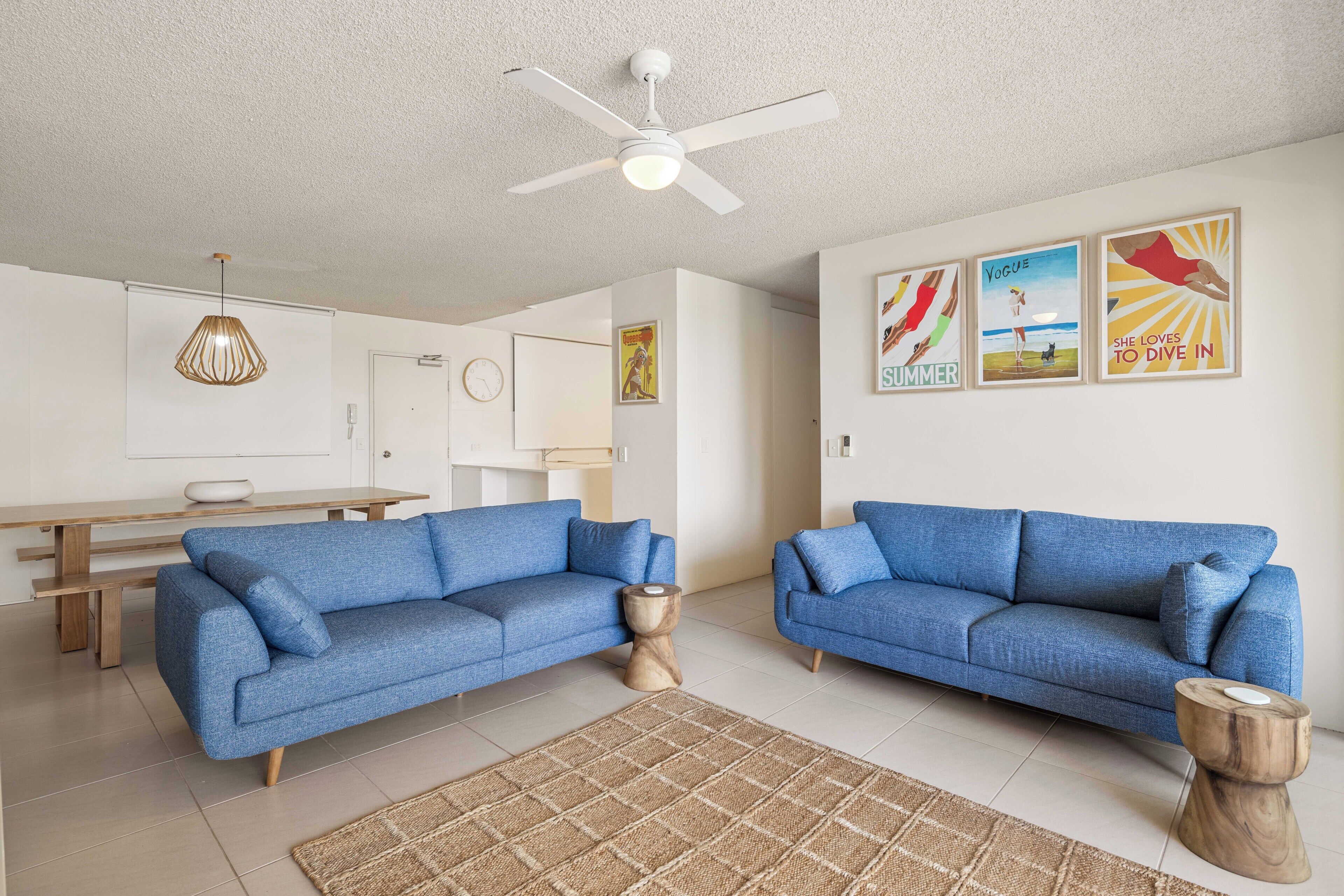 A modern living room with two blue sofas, a wooden coffee table, a ceiling fan, and wall art.