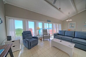 A spacious living room with a large window, a grey sofa, a white coffee table, and a ceiling fan. - Ocean Front 3 bed 3 bath sleeps 12 & Pet Friendly (Panama City Beach)