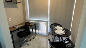 A small, modern room with a desk, two chairs, a table set for two, and a large window with blinds. - Studio next to Giovanni Metro Station | Vibra | Morumbi Stadium (São Paulo)