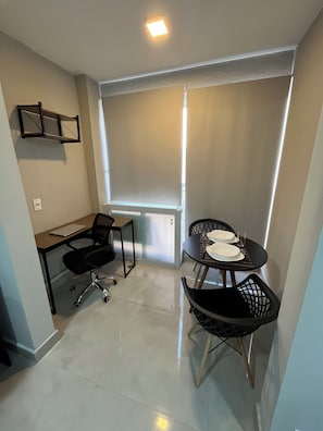 A small, modern room with a desk, two chairs, a table set for two, and a large window with blinds. - Studio next to Giovanni Metro Station | Vibra | Morumbi Stadium (São Paulo)