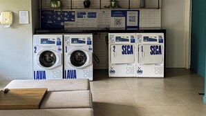 A laundry area with two washing machines labeled 'LAVA' and 'SECA,' a wall-mounted shelf with plants, and a bulletin board with informational posters. - Studio next to Giovanni Metro Station | Vibra | Morumbi Stadium (São Paulo)