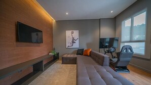 A modern living room with a sectional sofa, a flat-screen TV mounted on the wall, a large window, and a stationary exercise bike. - Studio next to Giovanni Metro Station | Vibra | Morumbi Stadium (São Paulo)