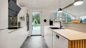 A modern kitchen with white cabinetry, a built-in dishwasher, and a large window overlooking a deck. - VillaVilla 944 - Blokhus, Nordjylland (Blokhus)