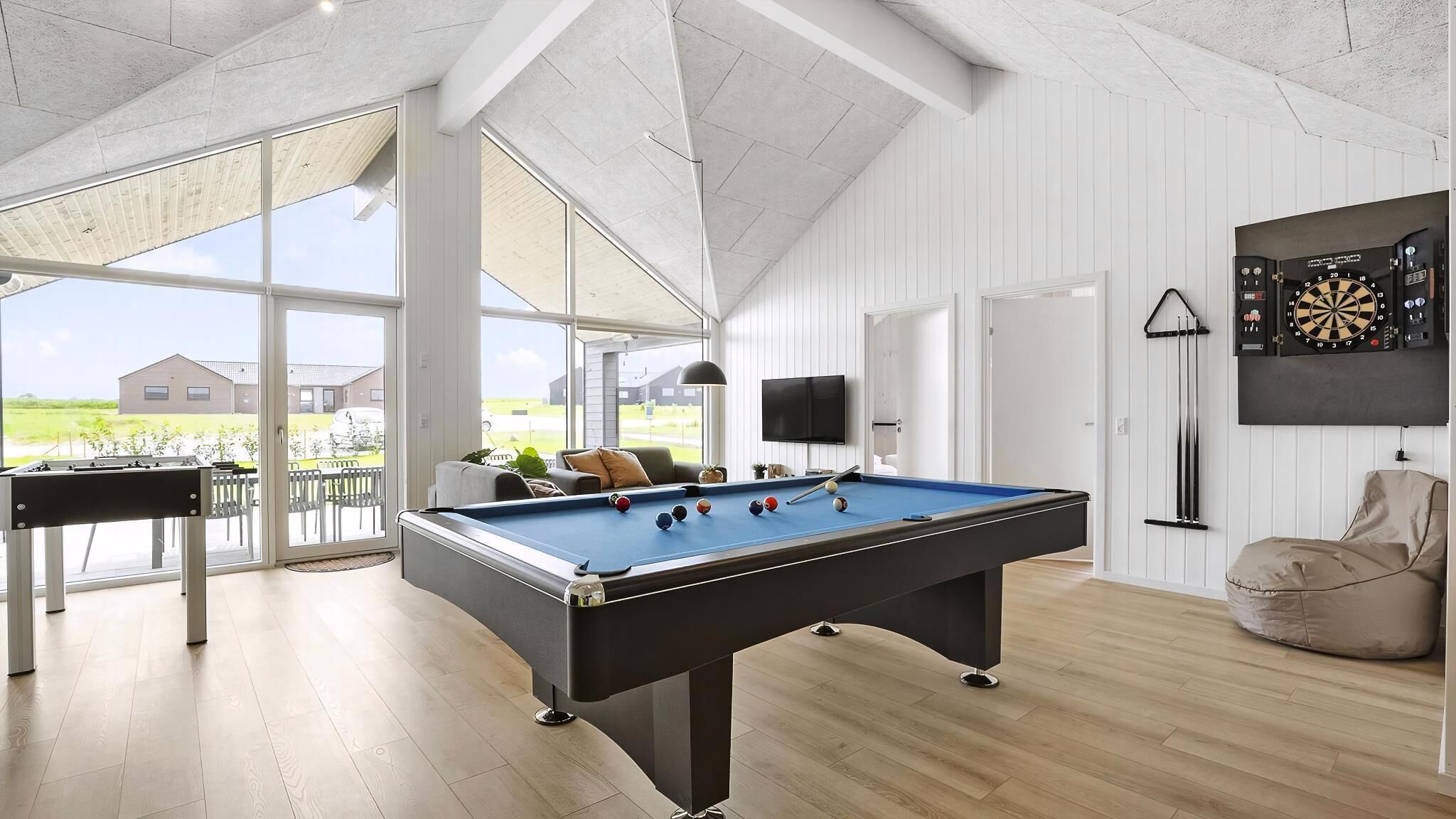 Ein Billardtisch mit Kugeln, eine Dartscheibe und ein Fernseher in einem Raum mit Holzböden und großen Fenstern.