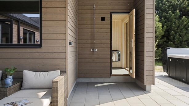Un patio exterior moderno con una pared de madera, un ventanal de vidrio que da a una sauna y una zona de estar con cojines.