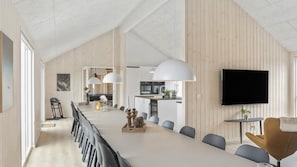 Un interior moderno con una larga mesa de comedor, varias sillas, una zona de cocina y un televisor en la pared.