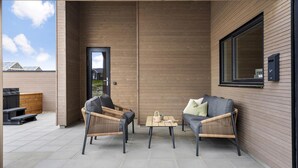 Un patio al aire libre con dos sillones de mimbre y una mesita, una pared de madera y una puerta que da a un área cubierta.