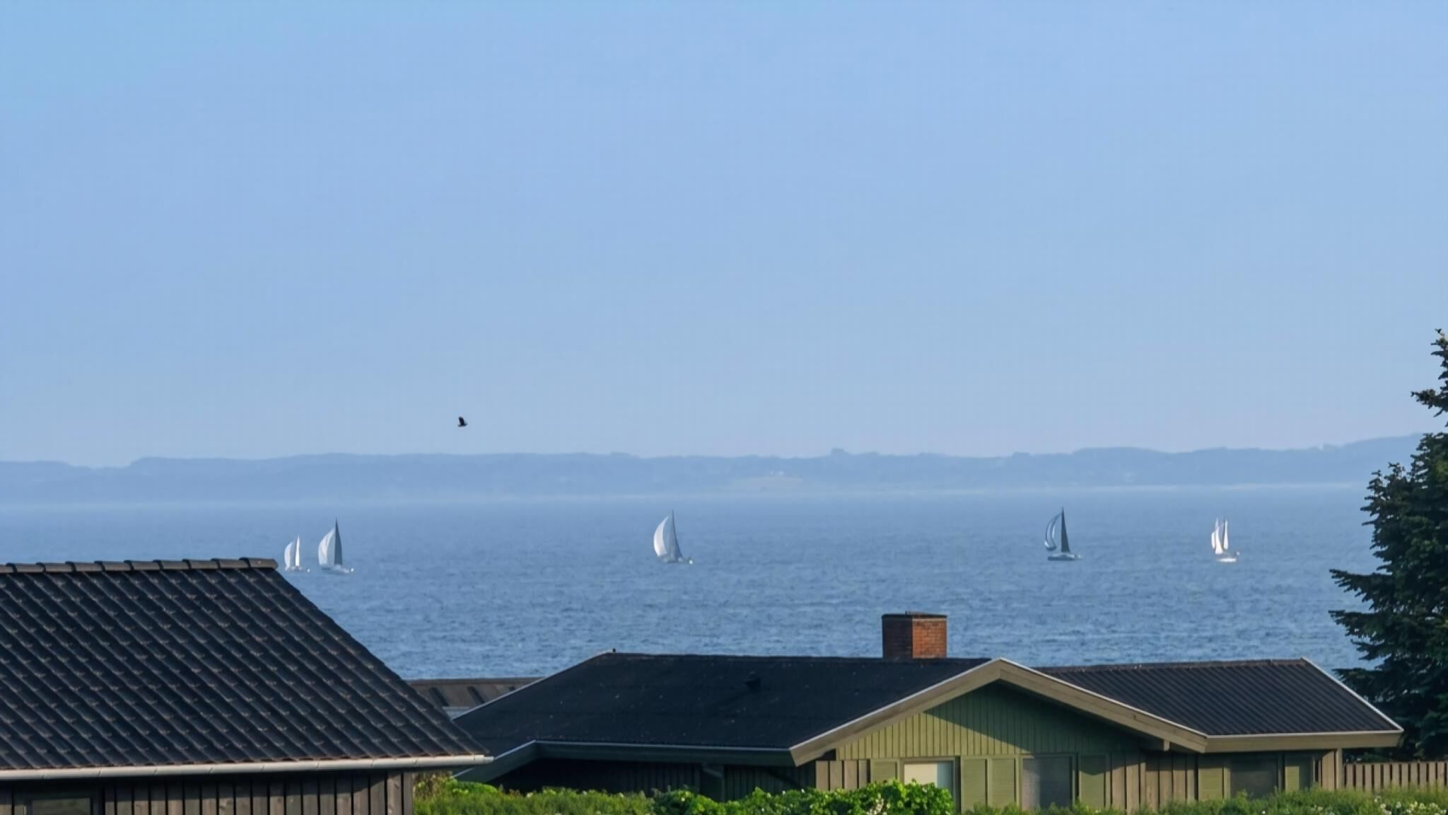 A coastal view with sailboats on the water, houses with sloped roofs, and a clear sky.