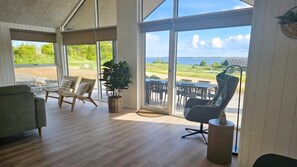 A spacious room with a large sliding glass door leading to an outdoor patio with a view of a grassy field and a body of water. - VillaVilla 922 - Faaborg, Fyn og øer (Faaborg)