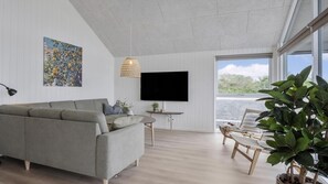 A modern living room with a grey sectional sofa, a flat-screen TV mounted on the wall, a small round table with a plant, and a large window offering a view of greenery. - VillaVilla 922 - Faaborg, Fyn og øer (Faaborg)