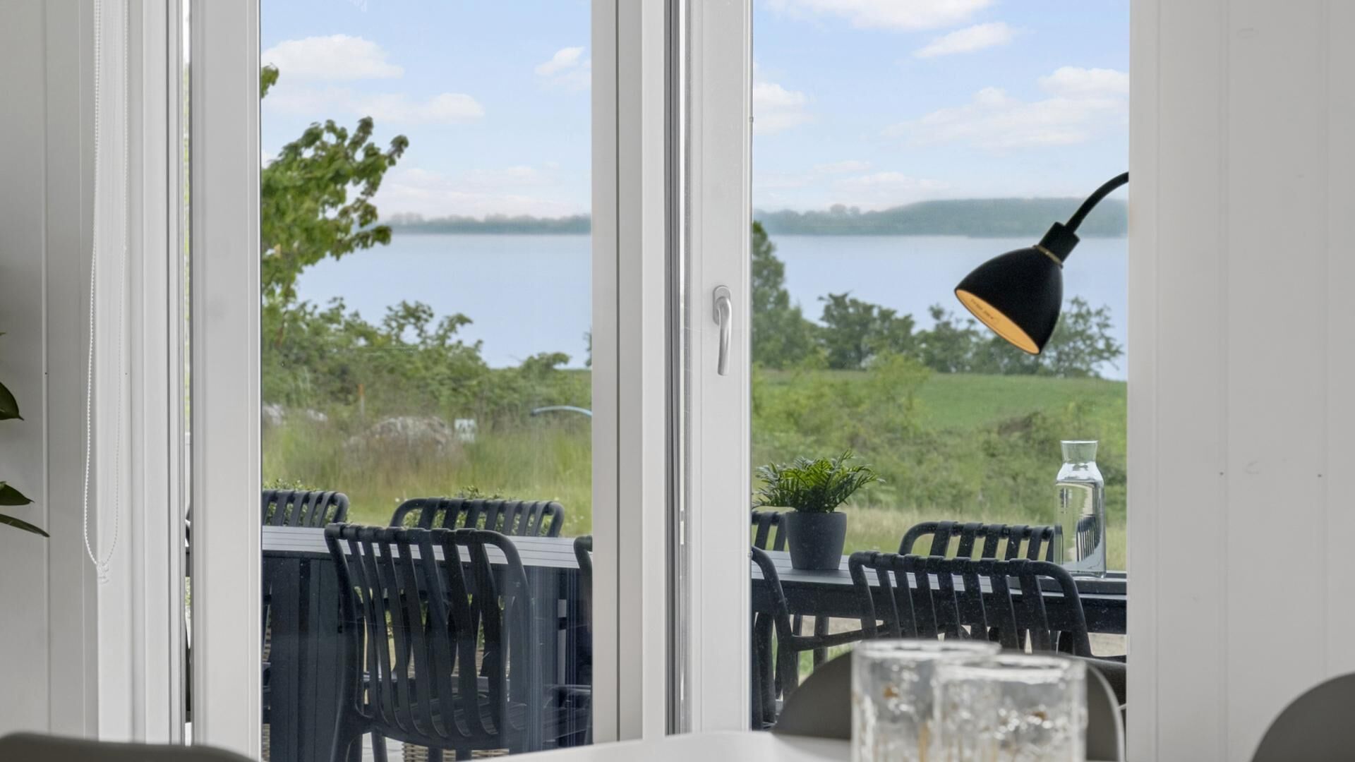 A room with a view of a lake, a table with chairs, a lamp, and a potted plant.