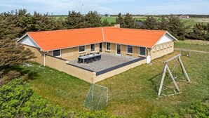 En stor, etetages bygning med rødt tag, en overdækket terrasse med bord og stole, og en indhegnet have.