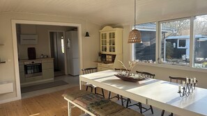 A modern kitchen with a central island, a dining area with a long table and benches, and a view of a house outside. - Cosy cottage 200 meters from the beach (Nykøbing Sj)