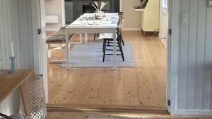 A room with a wooden floor, a dining table with chairs, a hanging light fixture, and a view into another room with a kitchen area. - Cosy cottage, 200 meters from the beach (Nykøbing Sj)
