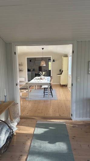 A room with a wooden floor, a dining table with chairs, a hanging light fixture, and a view into another room with a kitchen area. - Cosy cottage 200 meters from the beach (Nykøbing Sj)
