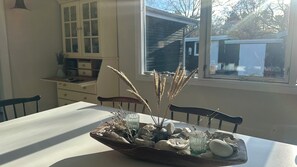 A dining table with a centerpiece of stones and dried plants, a white cabinet, and a window with a view of a house.